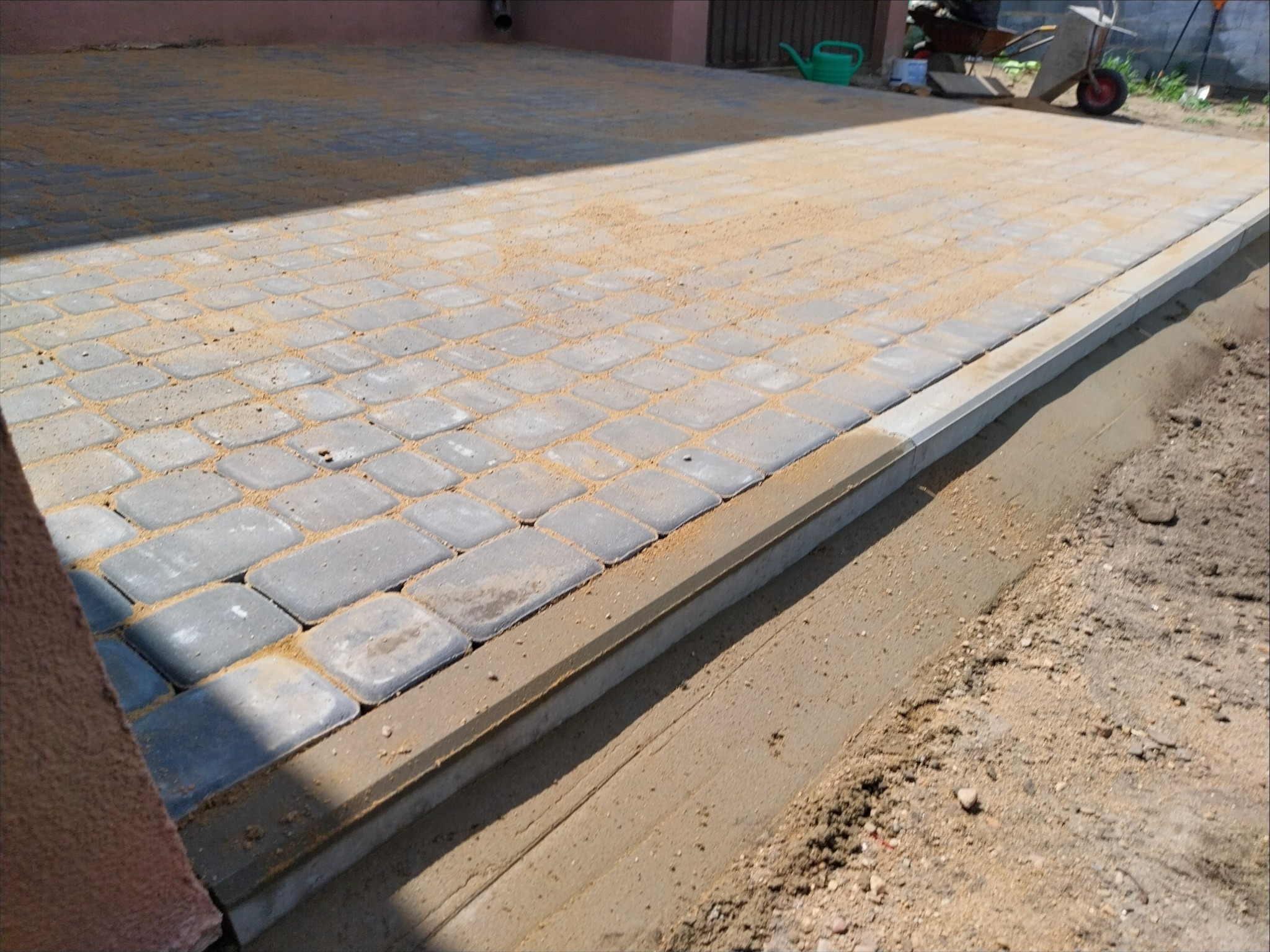 Szara kostka brukowa ułożona na podwórku, zasypana piaskiem, z betonową obwódką. W tle widoczne narzędzia ogrodnicze.
