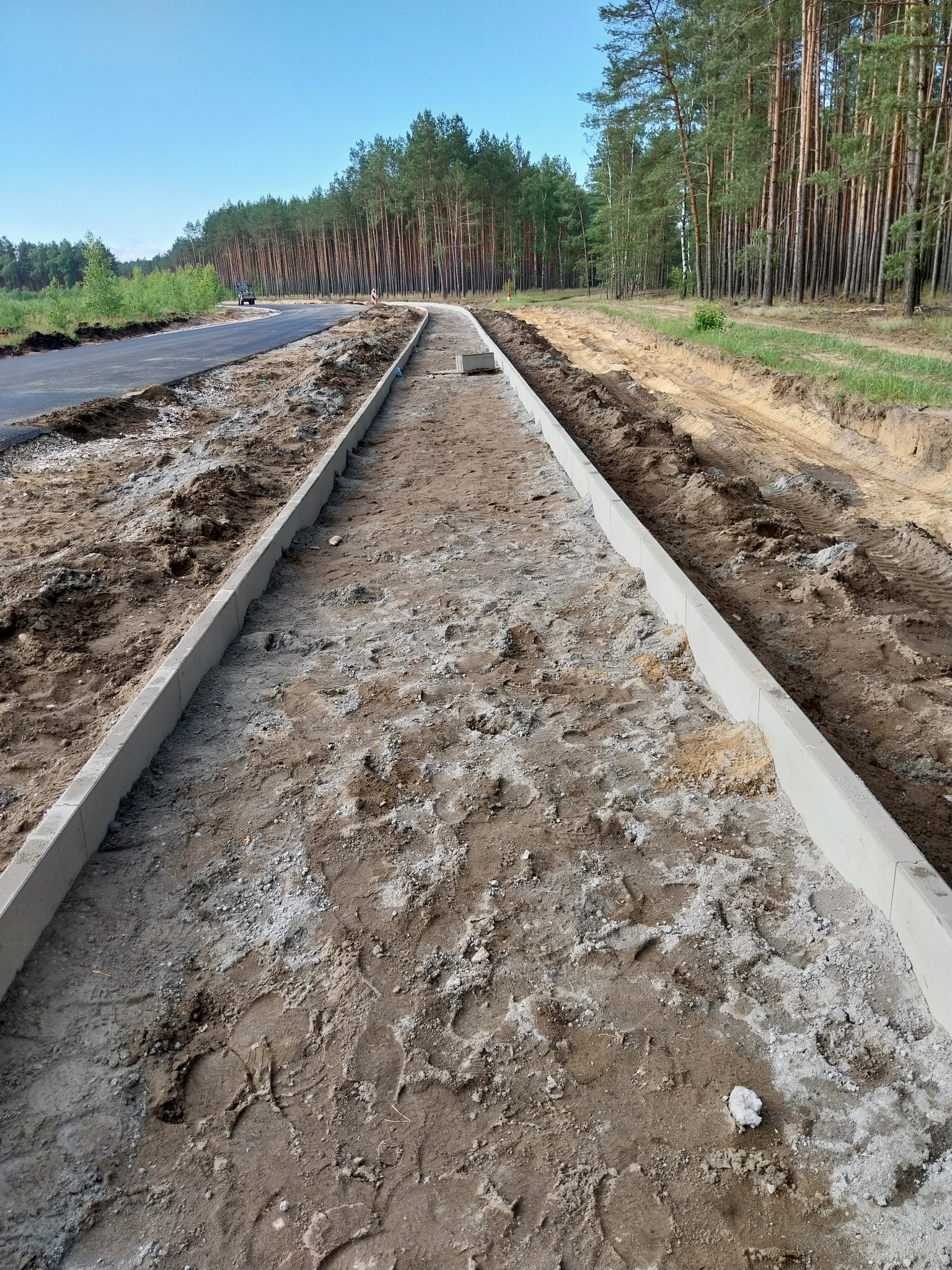 Przygotowany teren pod ścieżkę z obrzeżami z betonowych krawężników, widoczne ślady stóp w piasku, w tle droga asfaltowa i las.