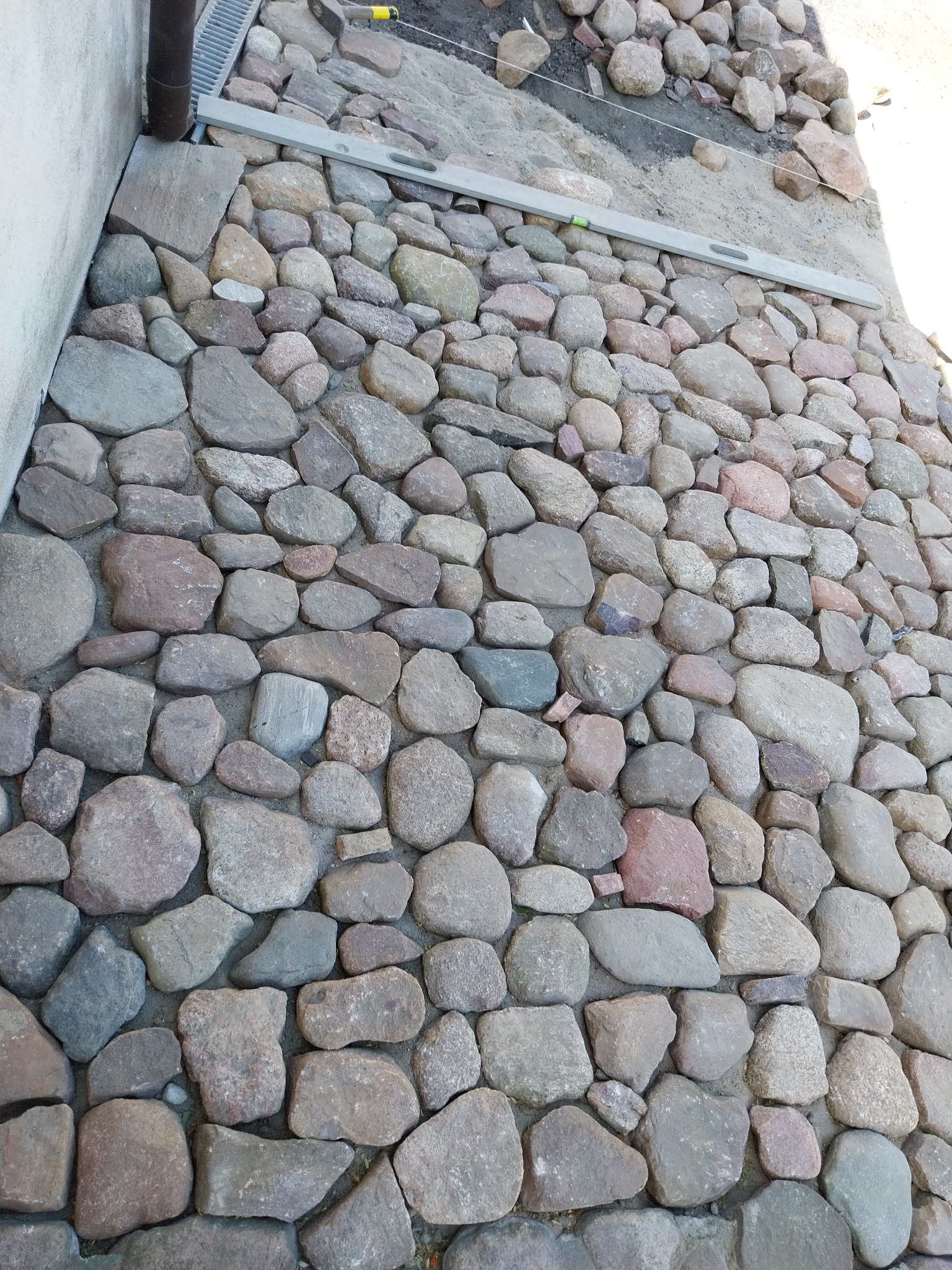 Nierówna nawierzchnia z nieregularnych kamieni polnych, poziomica i odpływ liniowy w tle, etap prac brukarskich.