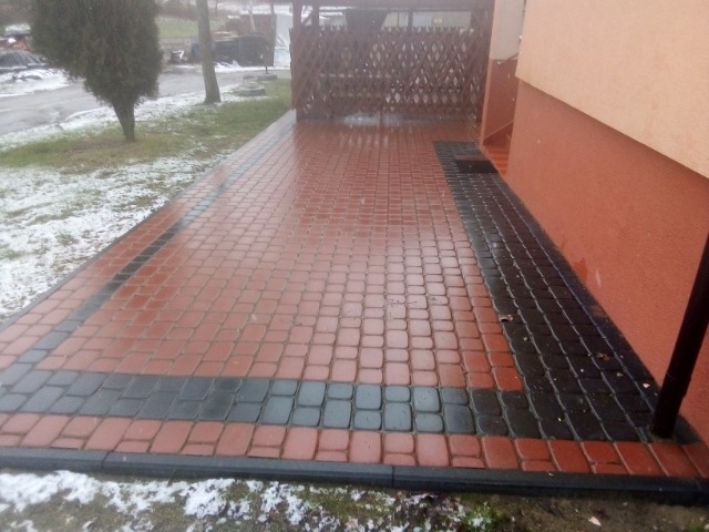 Czerwono-czarna kostka brukowa ułożona na podjeździe, widoczne ślady wilgoci i resztki śniegu.