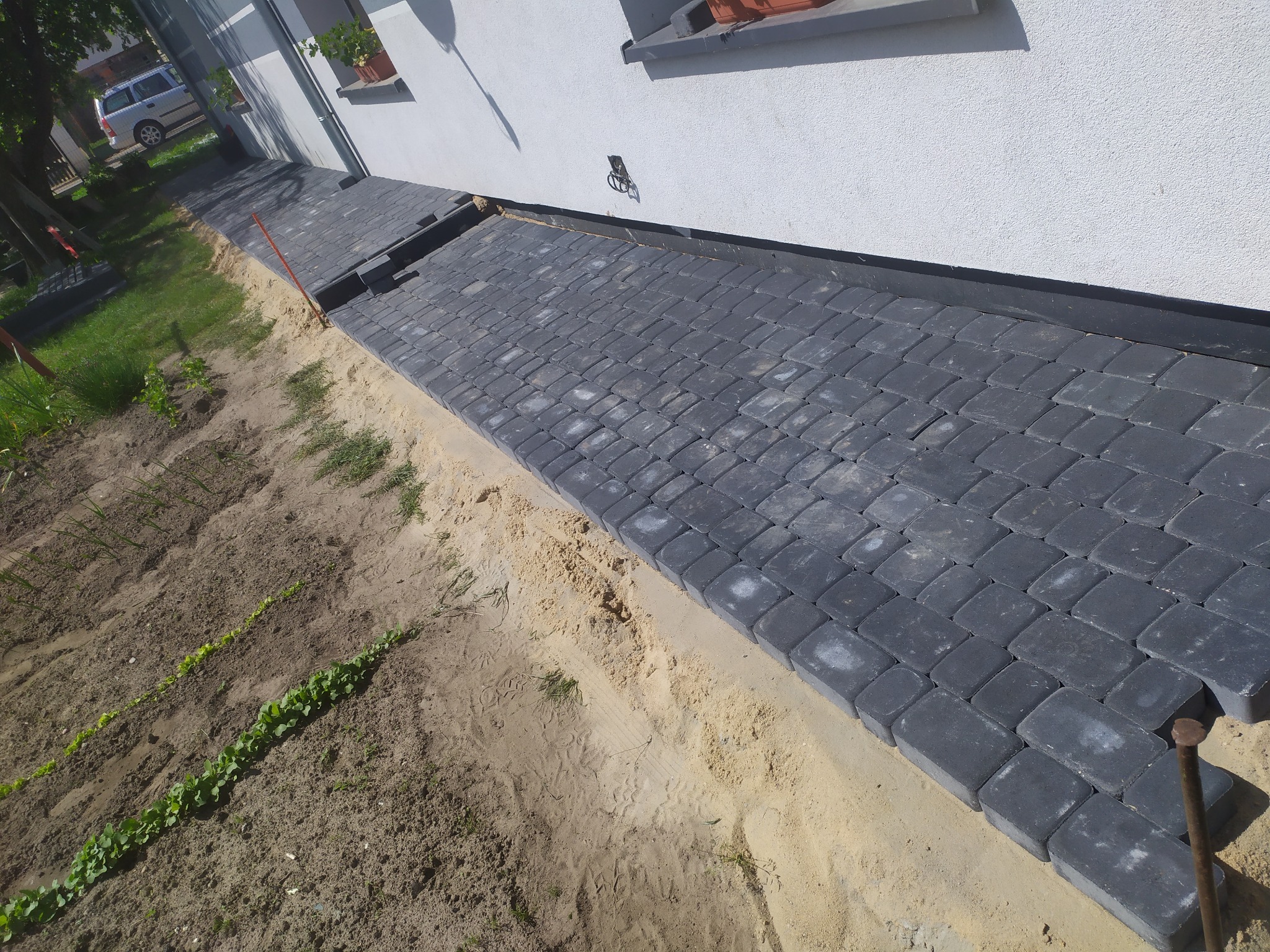 Świeżo ułożona, ciemnoszara kostka brukowa przy białej ścianie domu, z widoczną warstwą piasku i fragmentem ogrodu z młodymi roślinami.