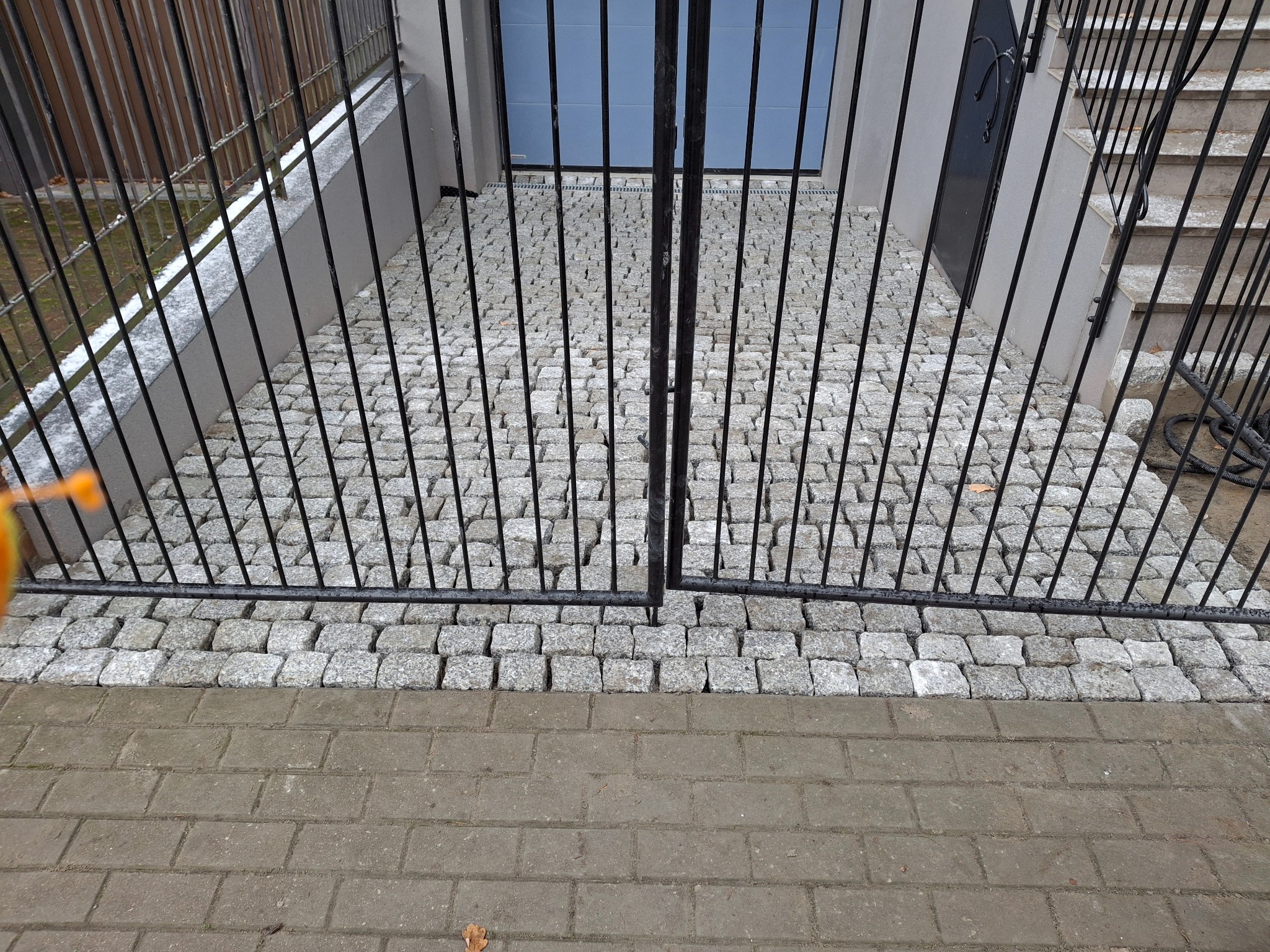 Granitowa kostka brukowa ułożona na schodach prowadzących do budynku, widoczna przez czarną, metalową bramę.