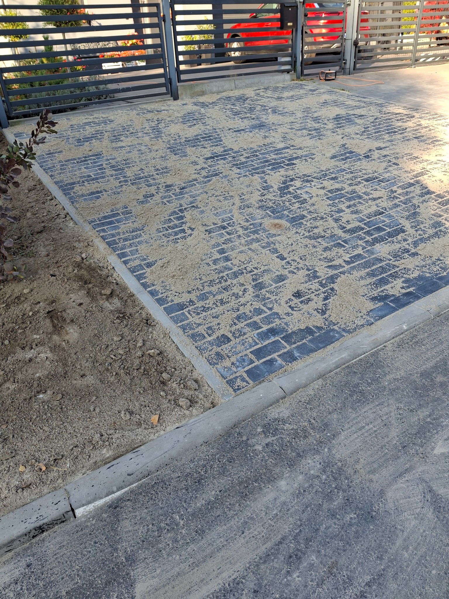 Nowo ułożona, czarna kostka brukowa zasypana piaskiem kwarcowym, widoczne obrzeża betonowe i fragment ogrodzenia posesji.