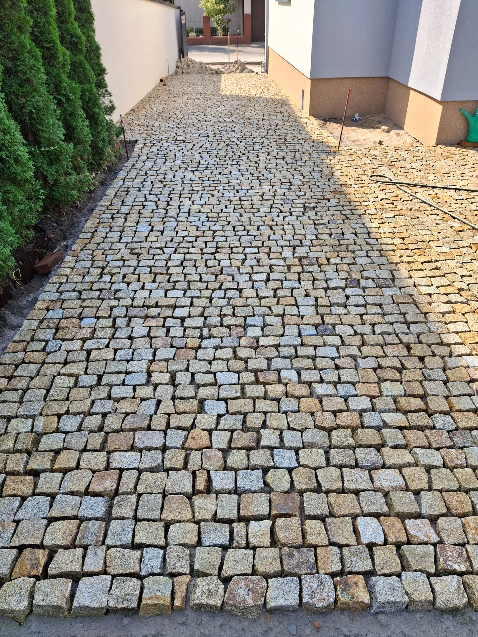 Ułożona droga z kostki brukowej w odcieniach szarości i żółci prowadząca do domu, widok z perspektywy osoby stojącej na początku drogi, z zielonymi krzewami po lewej stronie i domem po prawej stronie.