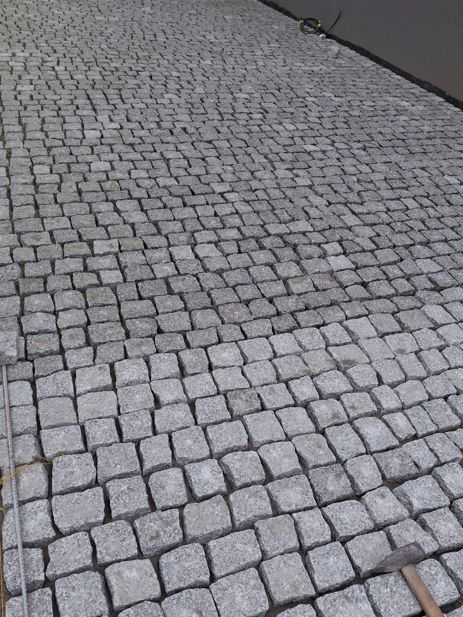 Szara kostka granitowa ułożona na piasku, widoczny fragment kabla i młotek brukarski.