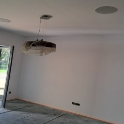 Łukasz Winowski - Wnętrze pokoju w trakcie malowania: ściany świeżo pomalowane na biało, podłoga zabezpieczona folią malarską, wisząca lampa przykryta folią ochronną, widoczne gniazdka elektryczne i otwór na głośnik...