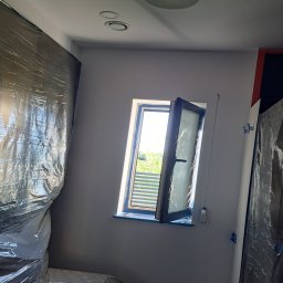 Łukasz Winowski - Pokój w trakcie prac wykończeniowych: ściany pomalowane na szaro, okno otwarte, zabezpieczone folią meble i ściany, widoczne ślady taśmy malarskiej.