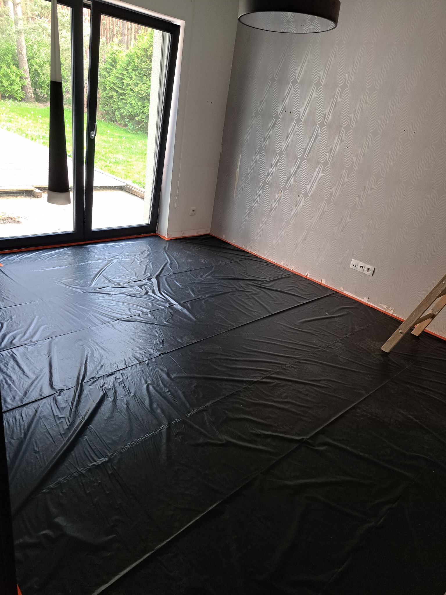 Pokój przygotowany do malowania: podłoga zabezpieczona czarną folią, ściany z wzorem, widoczna drabina i gniazdko elektryczne.