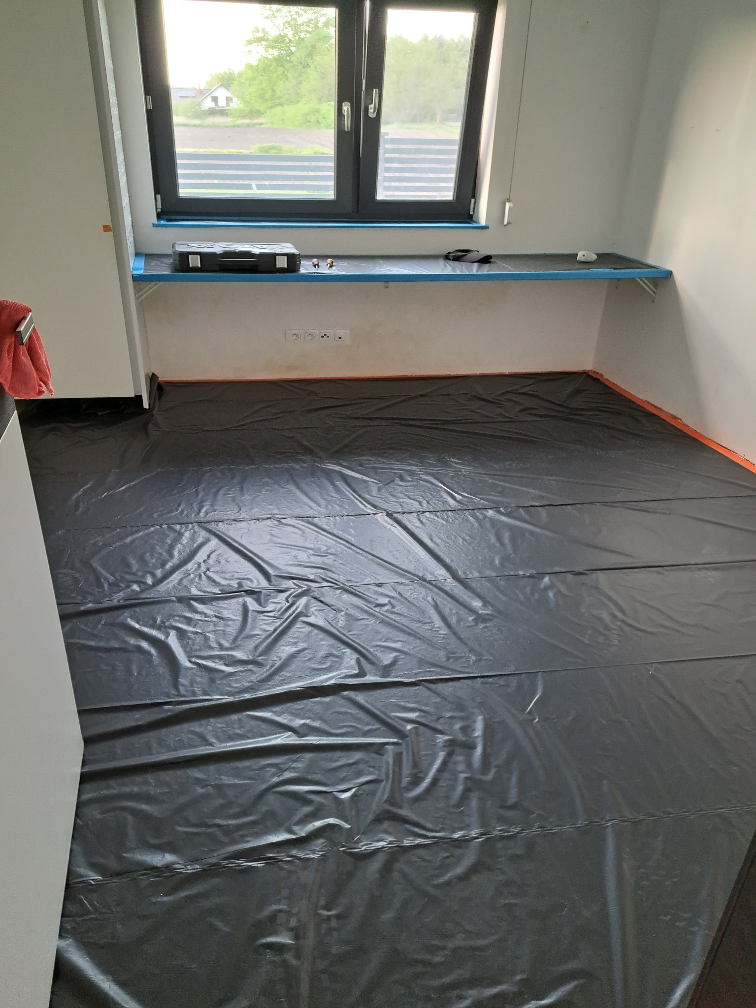 Przygotowanie do prac remontowych w pomieszczeniu: podłoga zabezpieczona czarną folią, okno z widokiem na zieleń, skrzynka z narzędziami na parapecie.