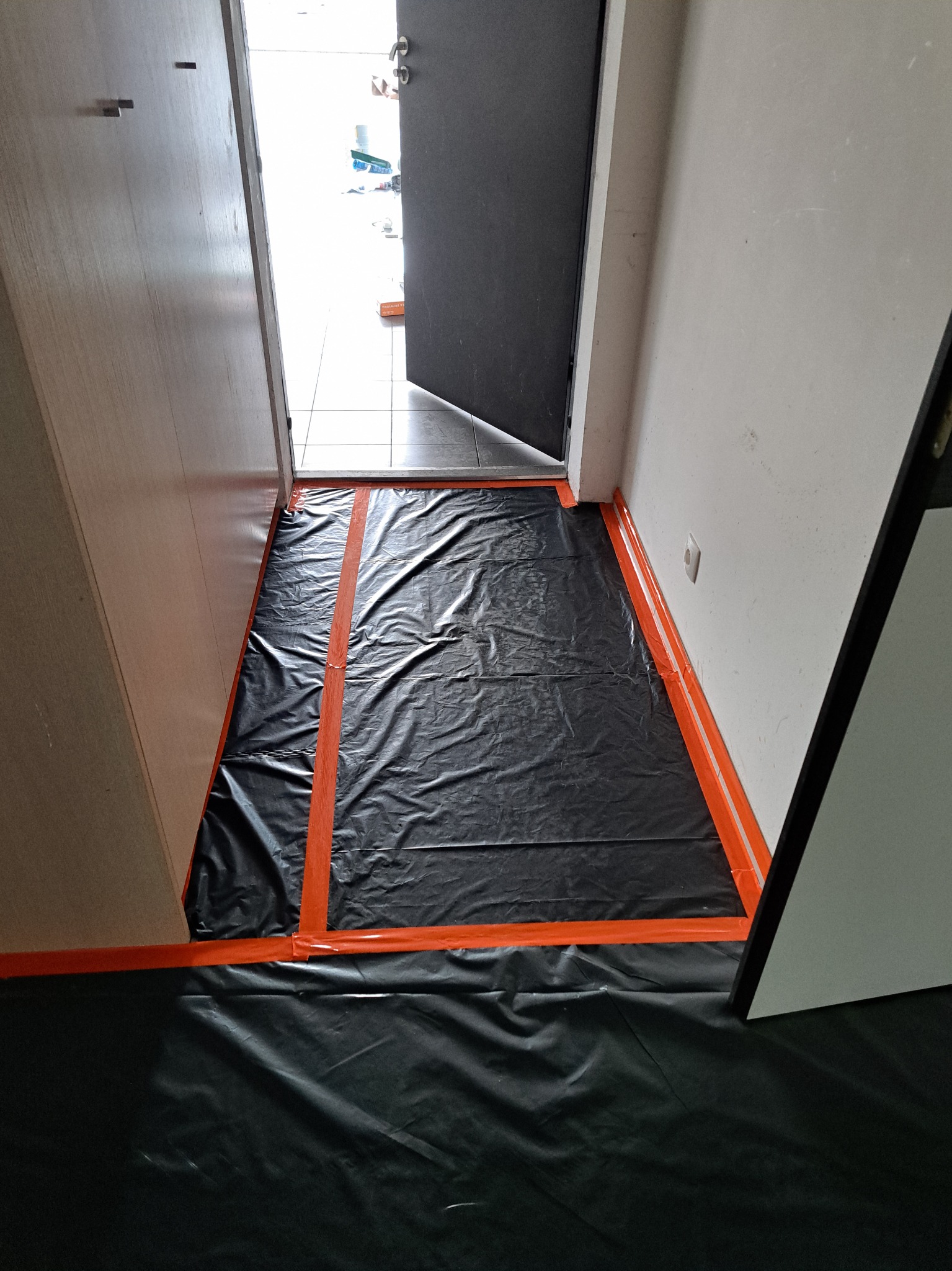 Przygotowanie podłoża przed pracami wykończeniowymi w korytarzu: podłoga i ściany zabezpieczone czarną folią i pomarańczową taśmą malarską.