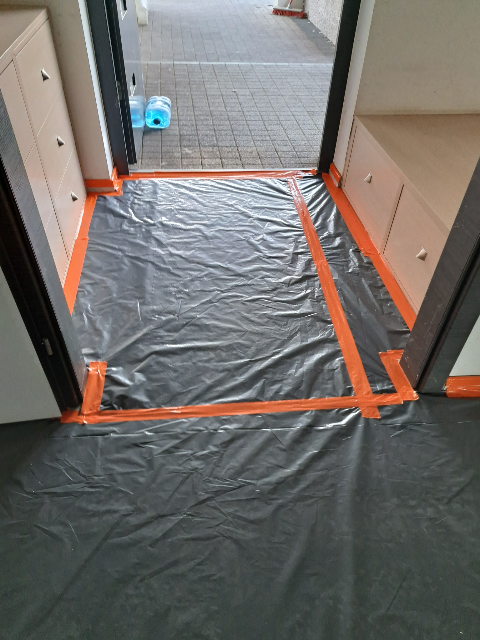 Przygotowanie do prac remontowych: podłoga w pokoju zabezpieczona czarną folią i pomarańczową taśmą malarską, widoczne szafki w zabudowie i fragment chodnika za progiem.