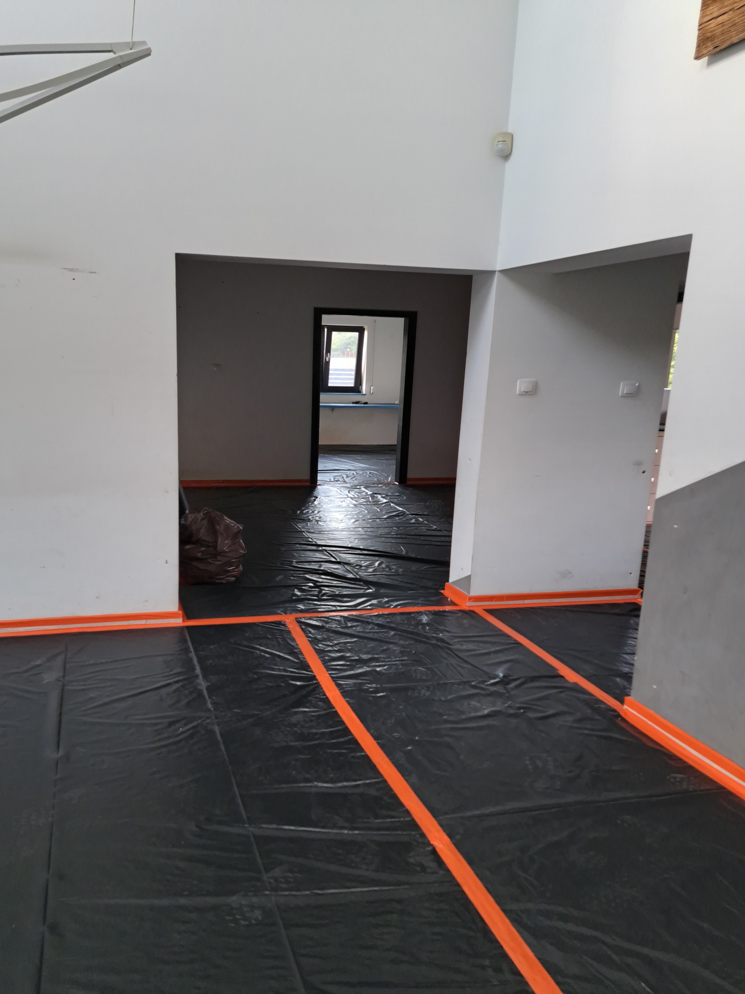Przygotowanie do prac wykończeniowych: zabezpieczona folią podłoga w pomieszczeniu z białymi ścianami, widoczna taśma malarska w kolorze pomarańczowym wzdłuż ścian i przejść, oraz worek z odpadami.