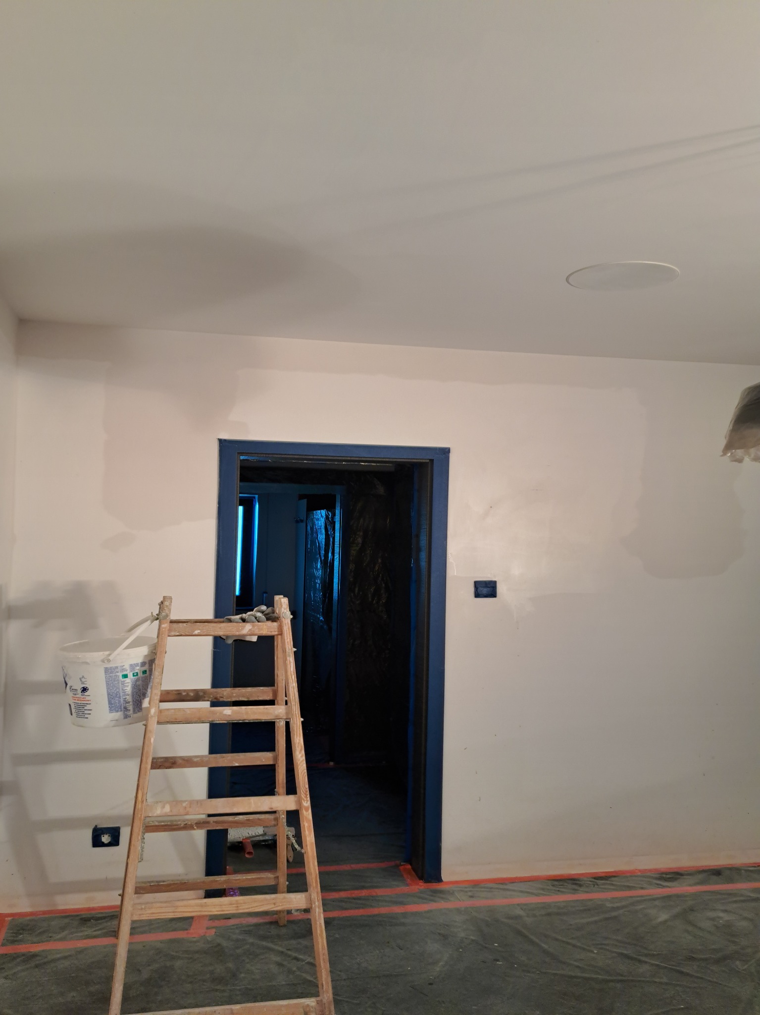 Pokój w trakcie malowania: drabina oparta o ścianę, wiadro z farbą, zabezpieczona folią podłoga, ściany z widocznymi śladami szpachlowania, rama drzwi pomalowana na niebiesko.