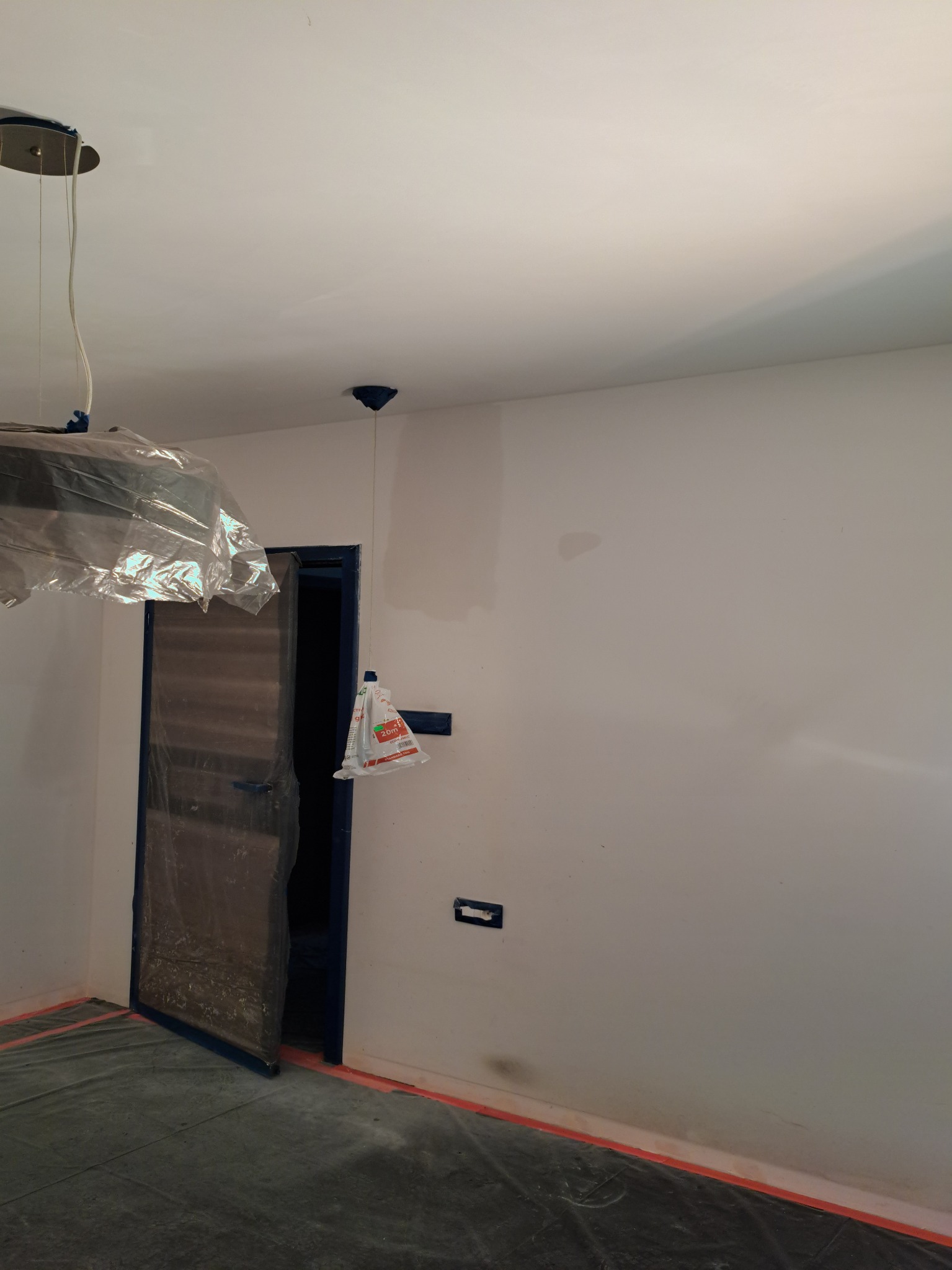 Przygotowane do malowania wnętrze: zabezpieczone folią meble i podłoga, ściany po szpachlowaniu, widoczne gniazdko elektryczne.