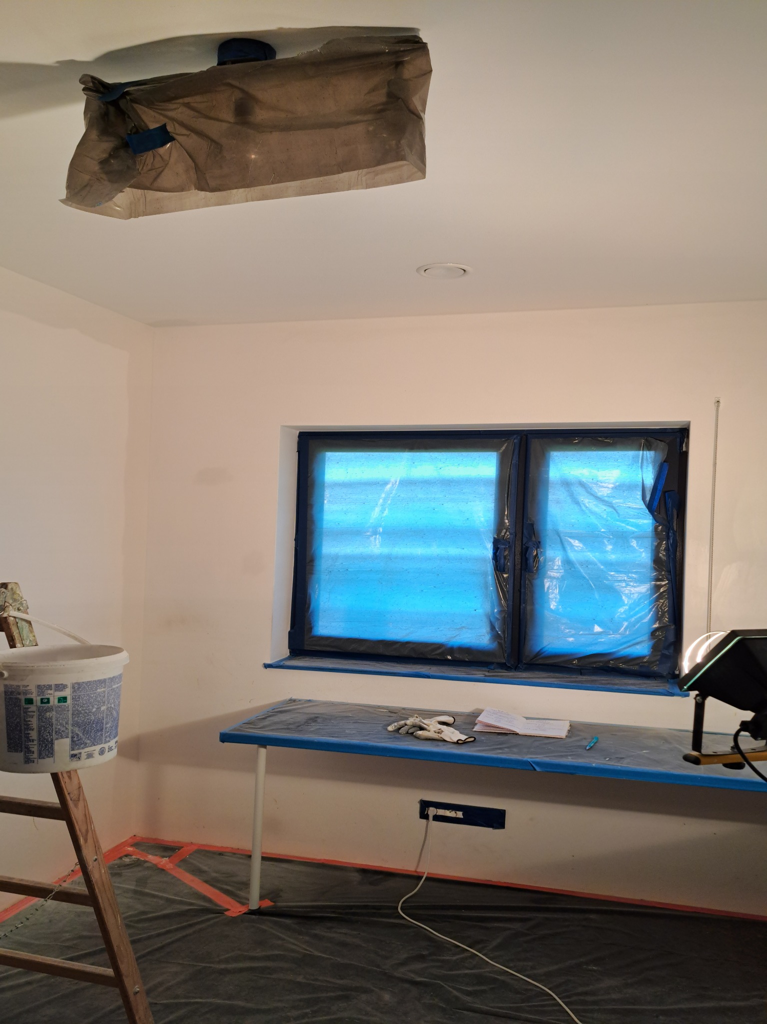 Przygotowane pomieszczenie do prac wykończeniowych: okno i meble zabezpieczone folią, podłoga przykryta, drabina z wiadrem farby w rogu, lampa oświetleniowa skierowana na ścianę.