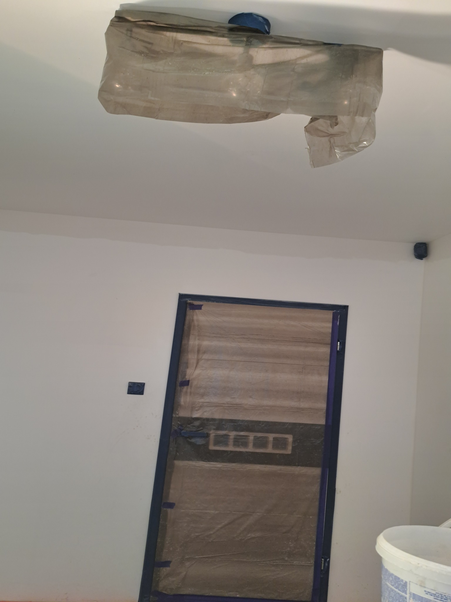 Przygotowane do malowania wnętrze: zabezpieczona folią lampa sufitowa i drzwi z niebieską futryną, ściany zagruntowane, widoczny fragment wiadra z farbą.