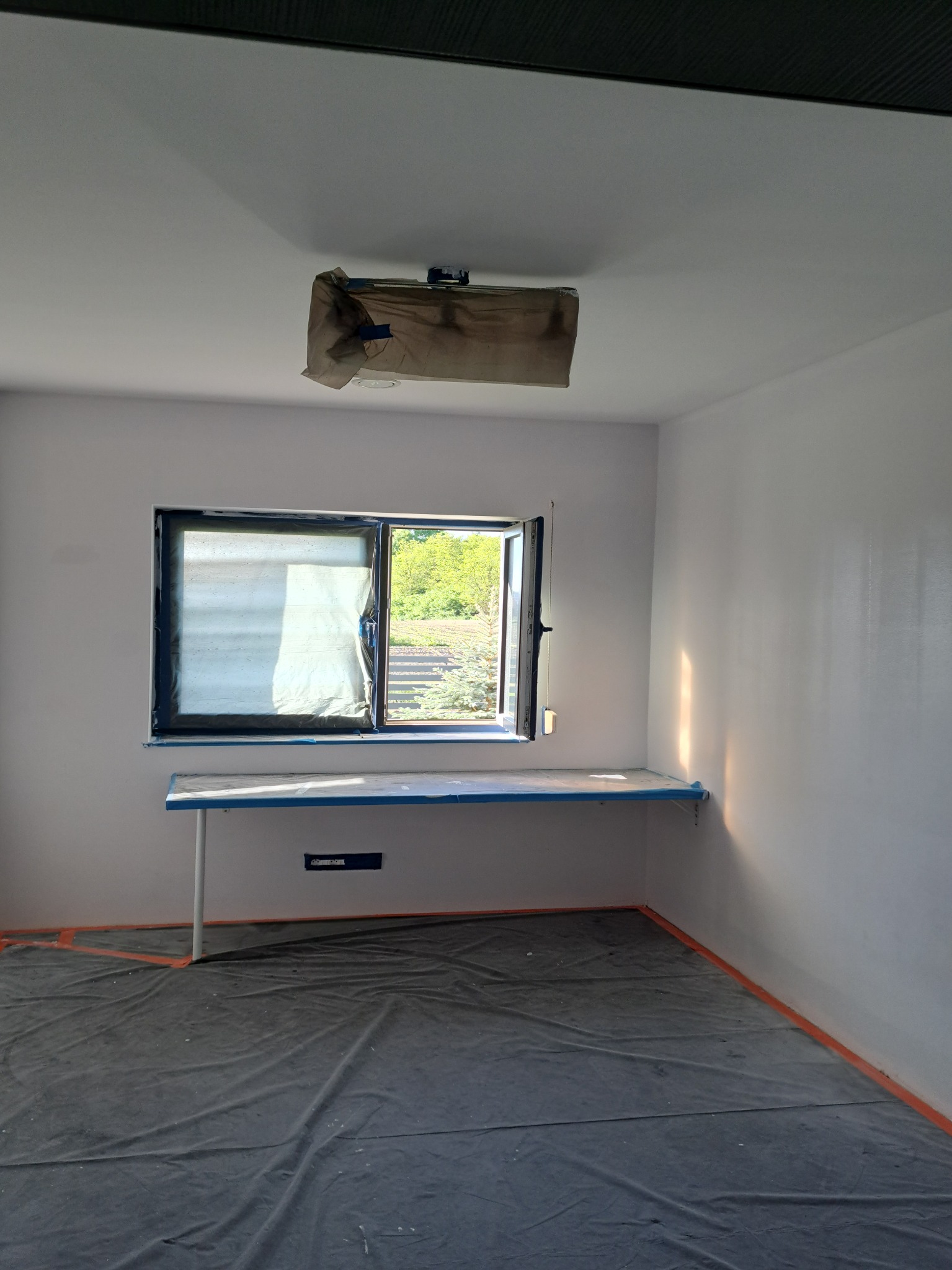 Pokój w trakcie prac wykończeniowych: świeżo pomalowane ściany na biało, okno zabezpieczone folią, podłoga przykryta folią ochronną, widoczny fragment zabezpieczonej lampy na suficie.