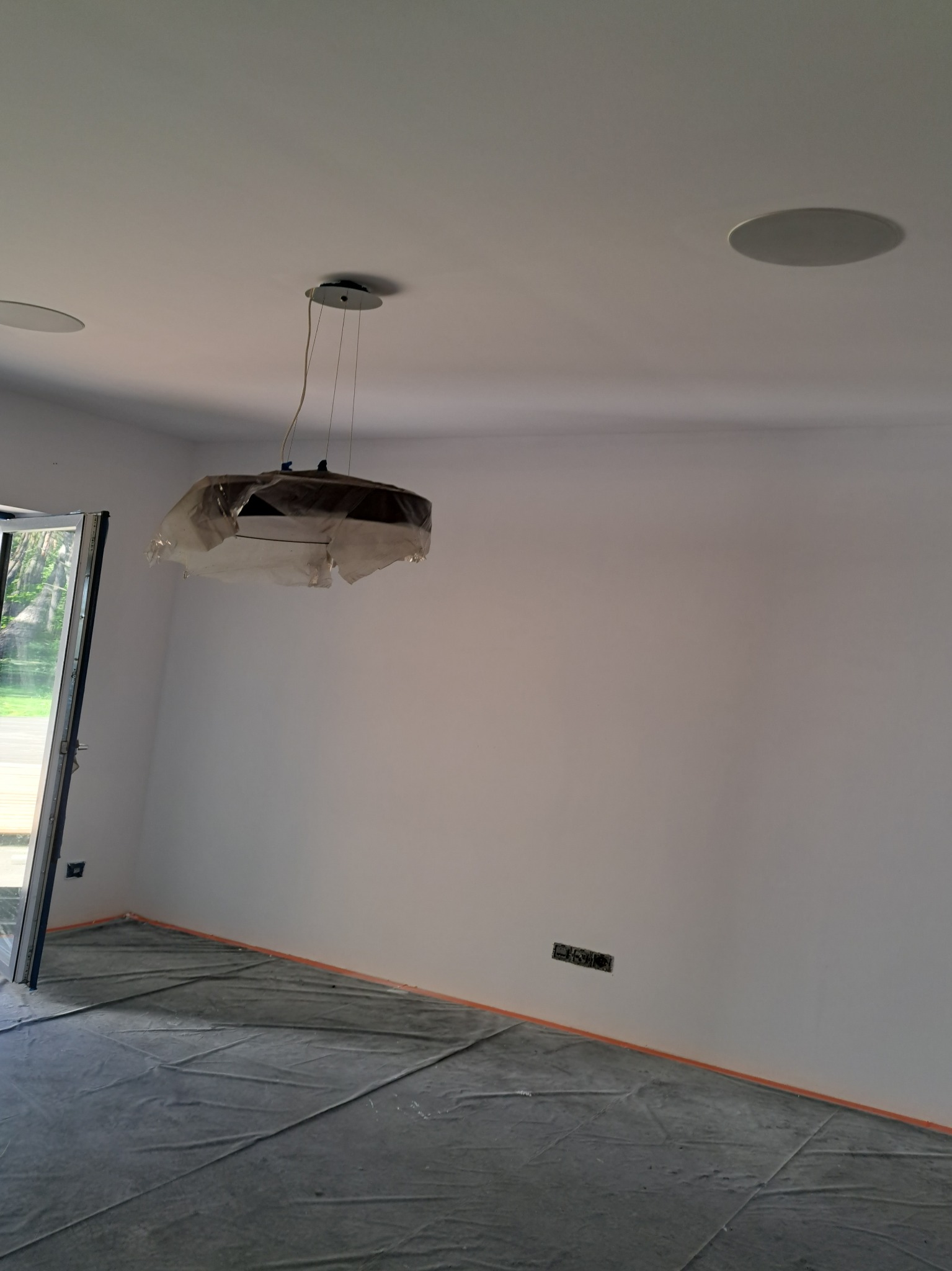 Wnętrze pokoju w trakcie malowania: ściany świeżo pomalowane na biało, podłoga zabezpieczona folią malarską, wisząca lampa przykryta folią ochronną, widoczne gniazdka elektryczne i otwór na głośnik...