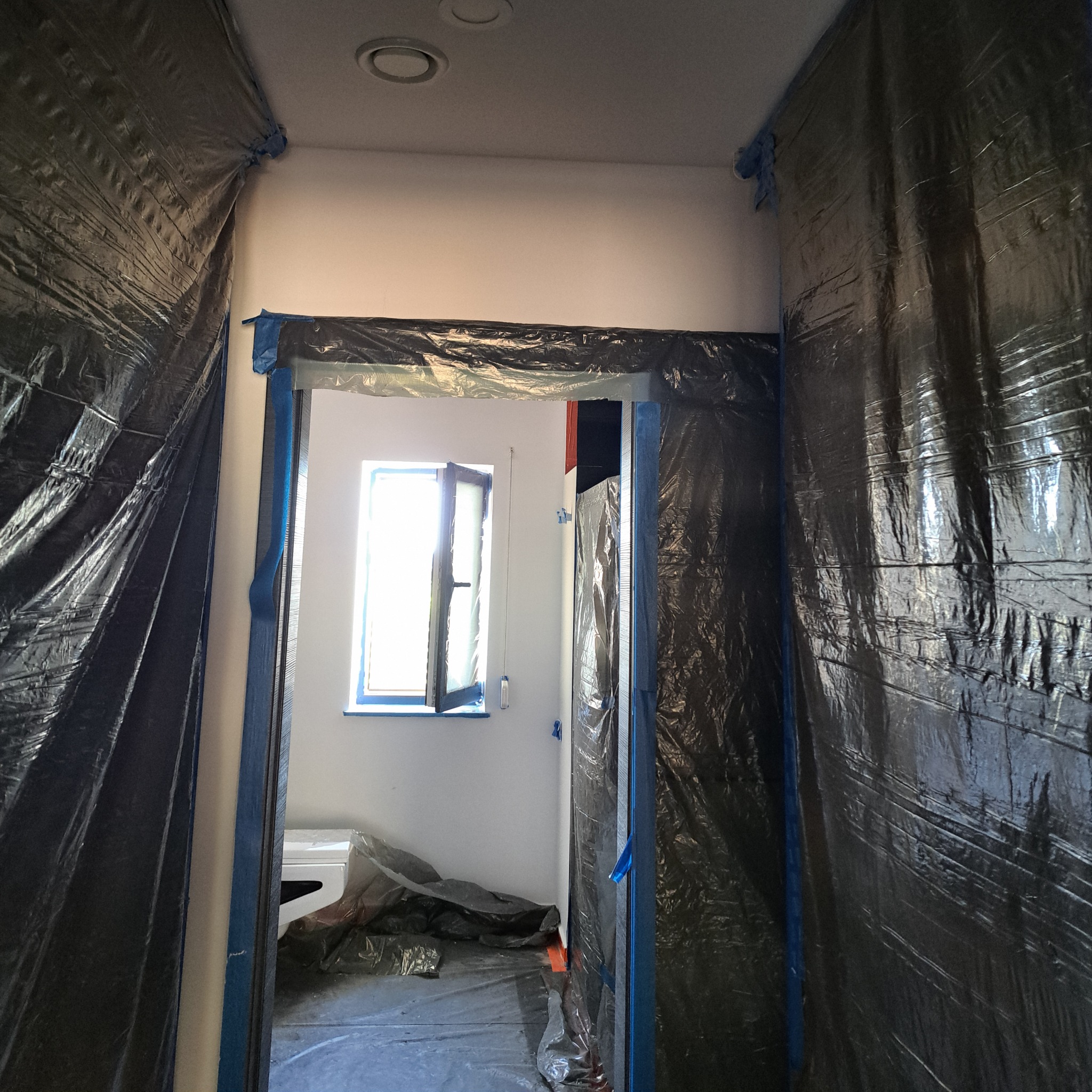 Przygotowanie pokoju do malowania: folia ochronna zabezpiecza ściany, podłogę i framugi okienne przed zabrudzeniem farbą, okno uchylone dla wentylacji.