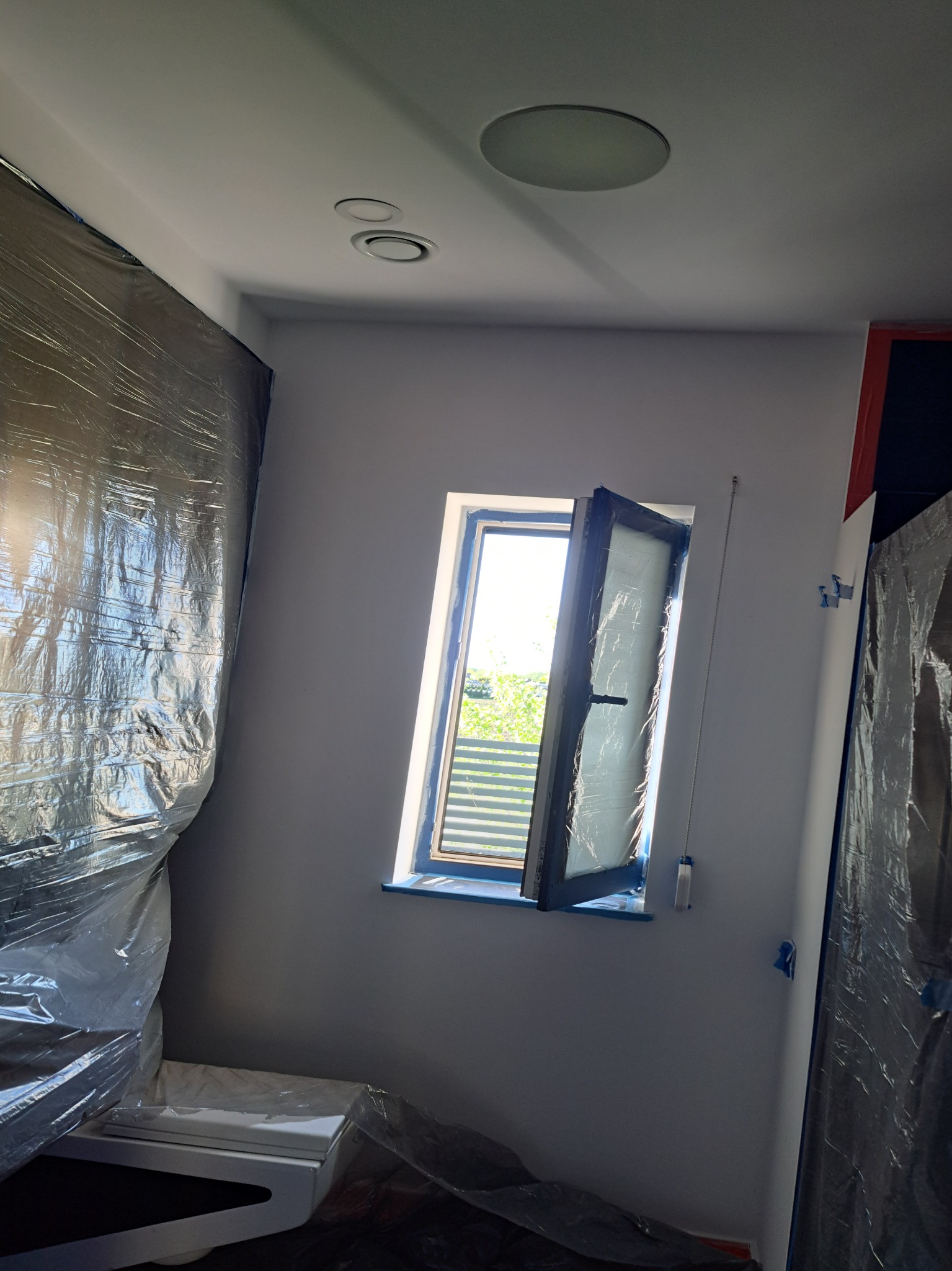 Pokój w trakcie prac wykończeniowych: ściany pomalowane na szaro, okno otwarte, zabezpieczone folią meble i ściany, widoczne ślady taśmy malarskiej.