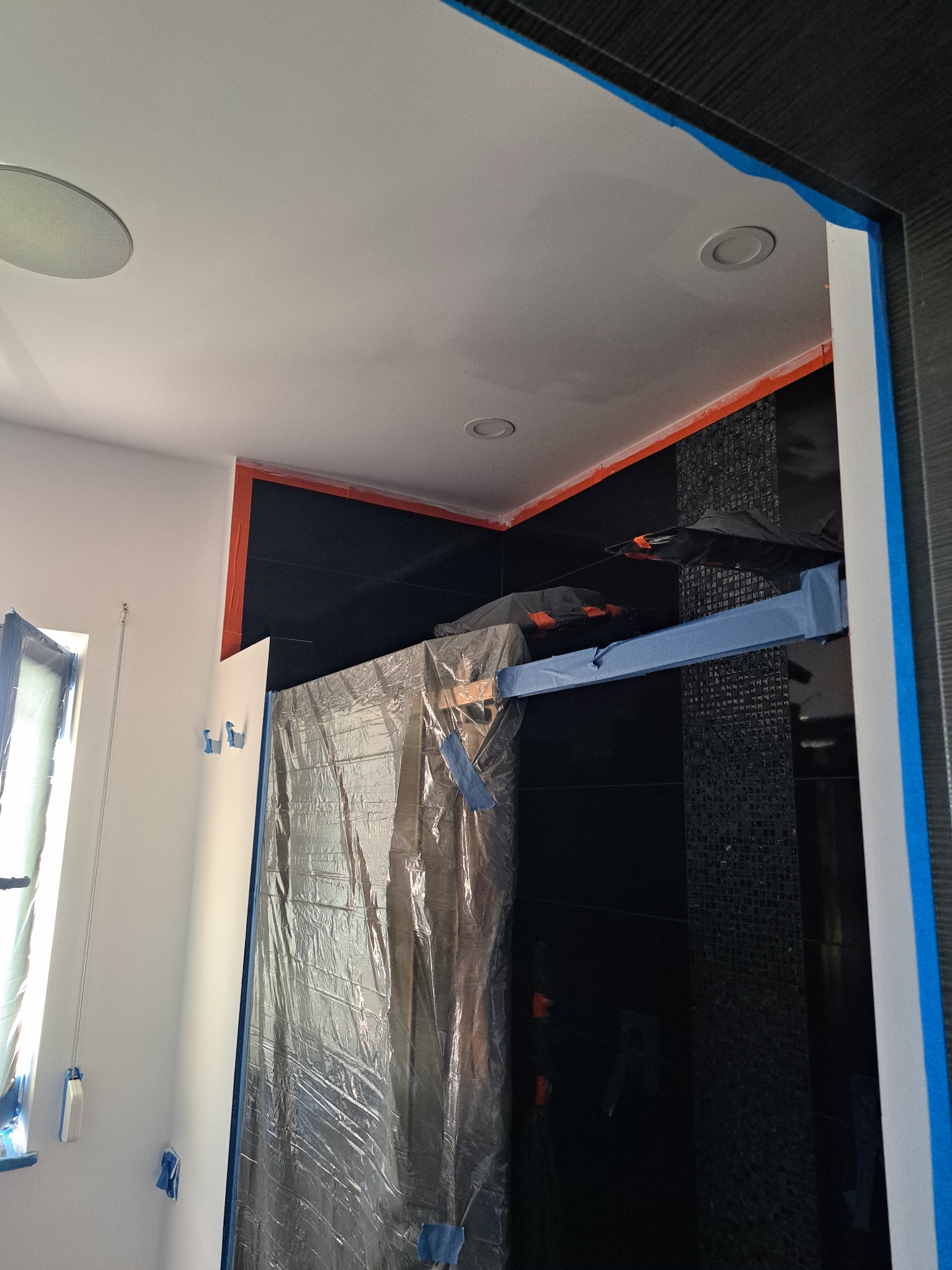 Łazienka w trakcie zabezpieczania folią i taśmą malarską przed pracami wykończeniowymi, widoczne czarne płytki mozaikowe i biała ściana z oknem.