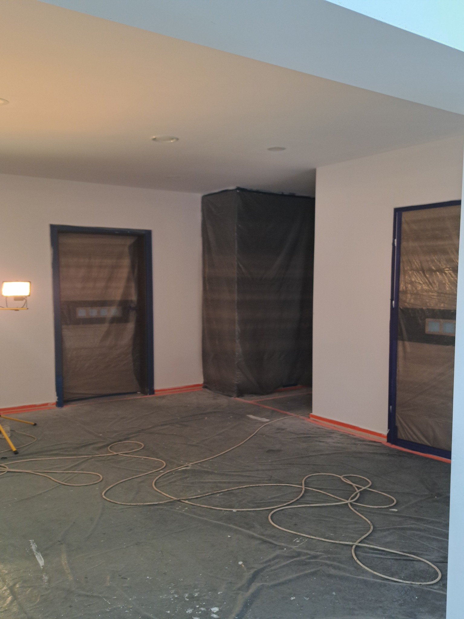 Przygotowane do malowania wnętrze: zakryte folią drzwi, podłoga i meble, ściany po szpachlowaniu, widoczny kabel lampy budowlanej.