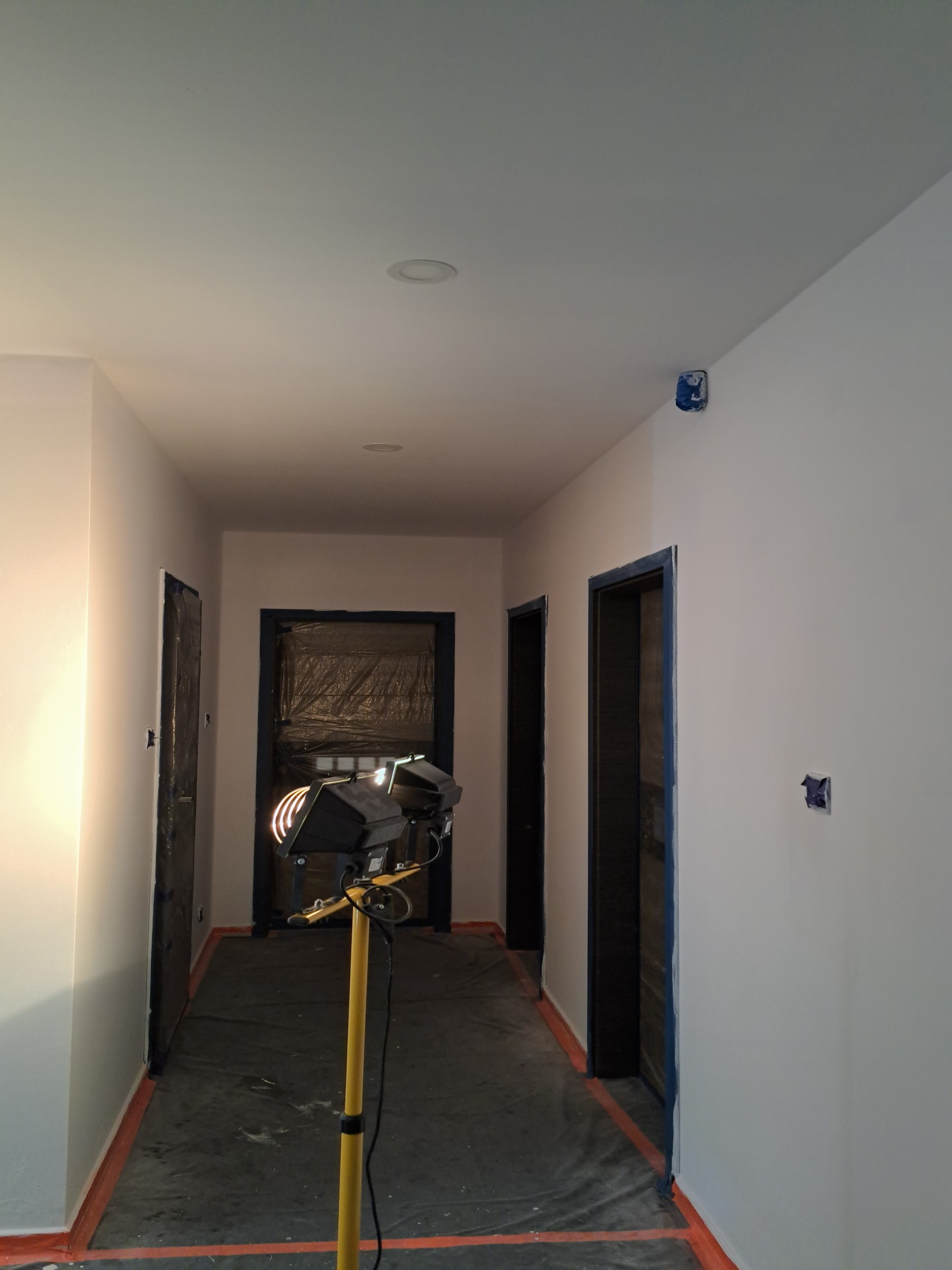 Korytarz w trakcie odświeżania: ściany pomalowane na biało, drzwi zabezpieczone folią i taśmą malarską, podłoga przykryta folią ochronną, widoczny halogen na statywie.