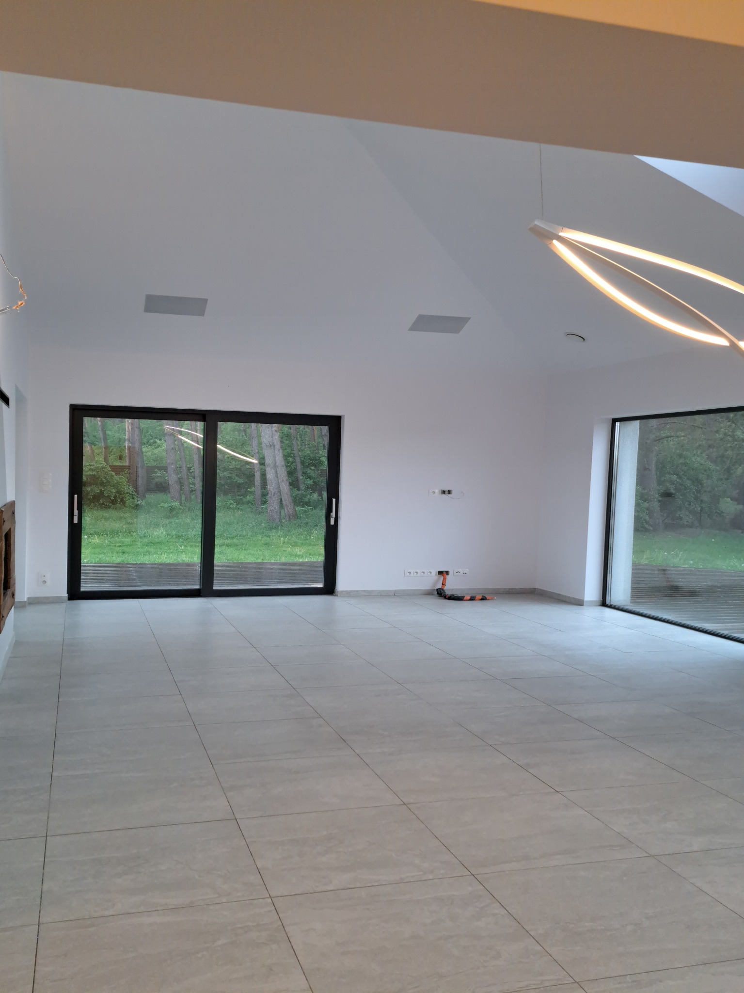 Wnętrze przestronnego, jasnego pokoju z dużymi przesuwnymi oknami wychodzącymi na zieleń, szare płytki na podłodze i designerska lampa sufitowa, widoczne gniazdka i przewody na ścianie.