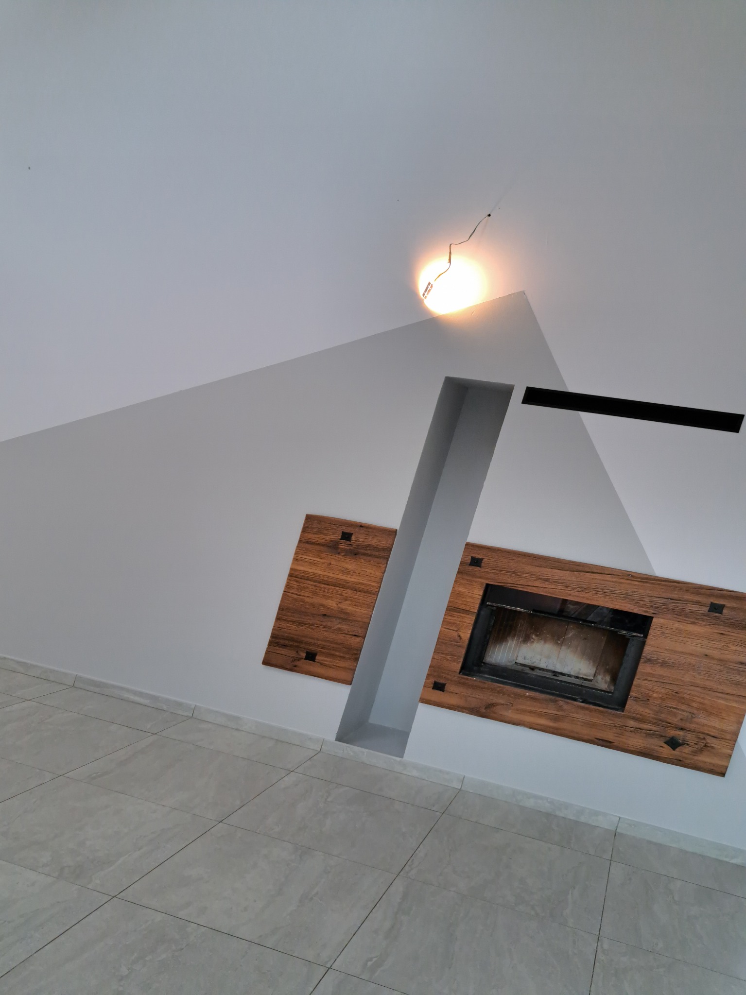 Nowoczesny kominek z drewnianą obudową wkomponowany w szarą ścianę i sufit, widok z dołu na jasną podłogę z płytek.