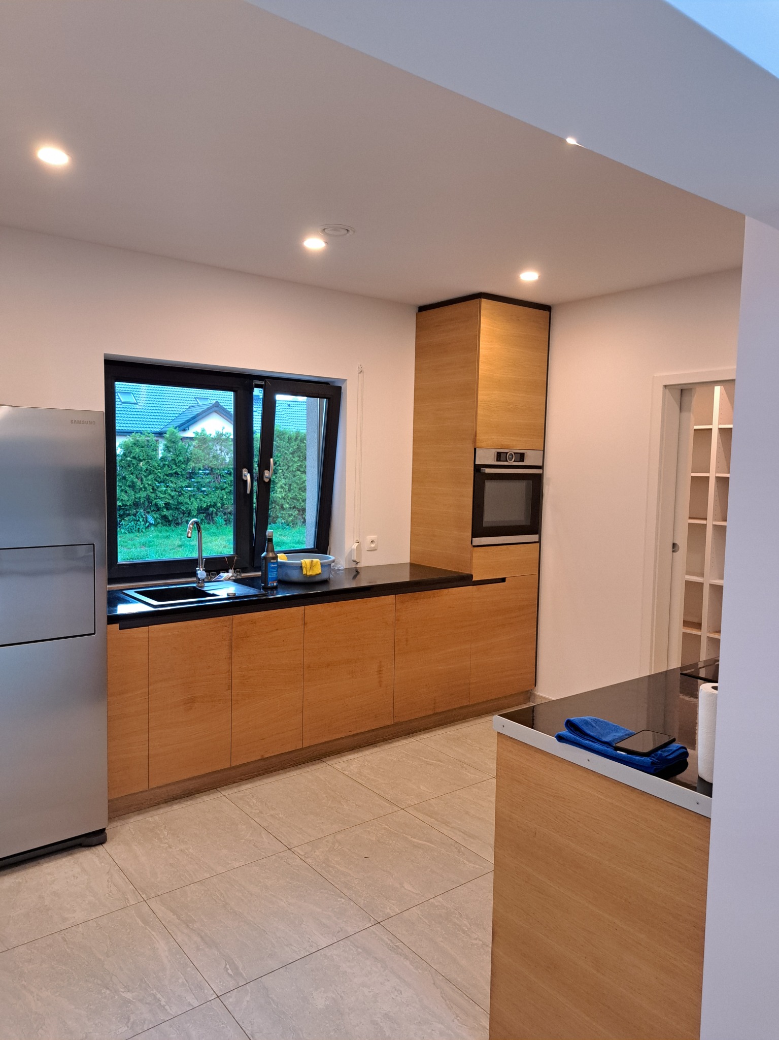 Jasna kuchnia z drewnianymi szafkami, czarnym blatem i srebrną lodówką Samsung. Widok na ogród przez okno nad zlewem. Na blacie przy oknie stoi butelka płynu do naczyń i miska z gąbką. Na wyspie...