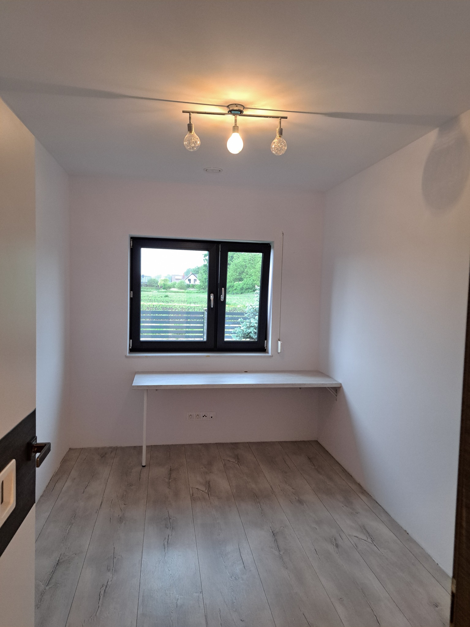 Wnętrze pokoju po odświeżeniu ścian na biało, z drewnianą podłogą, czarnym oknem z widokiem na ogród i zamontowanym blatem pod oknem, oświetlone lampą sufitową z trzema żarówkami.