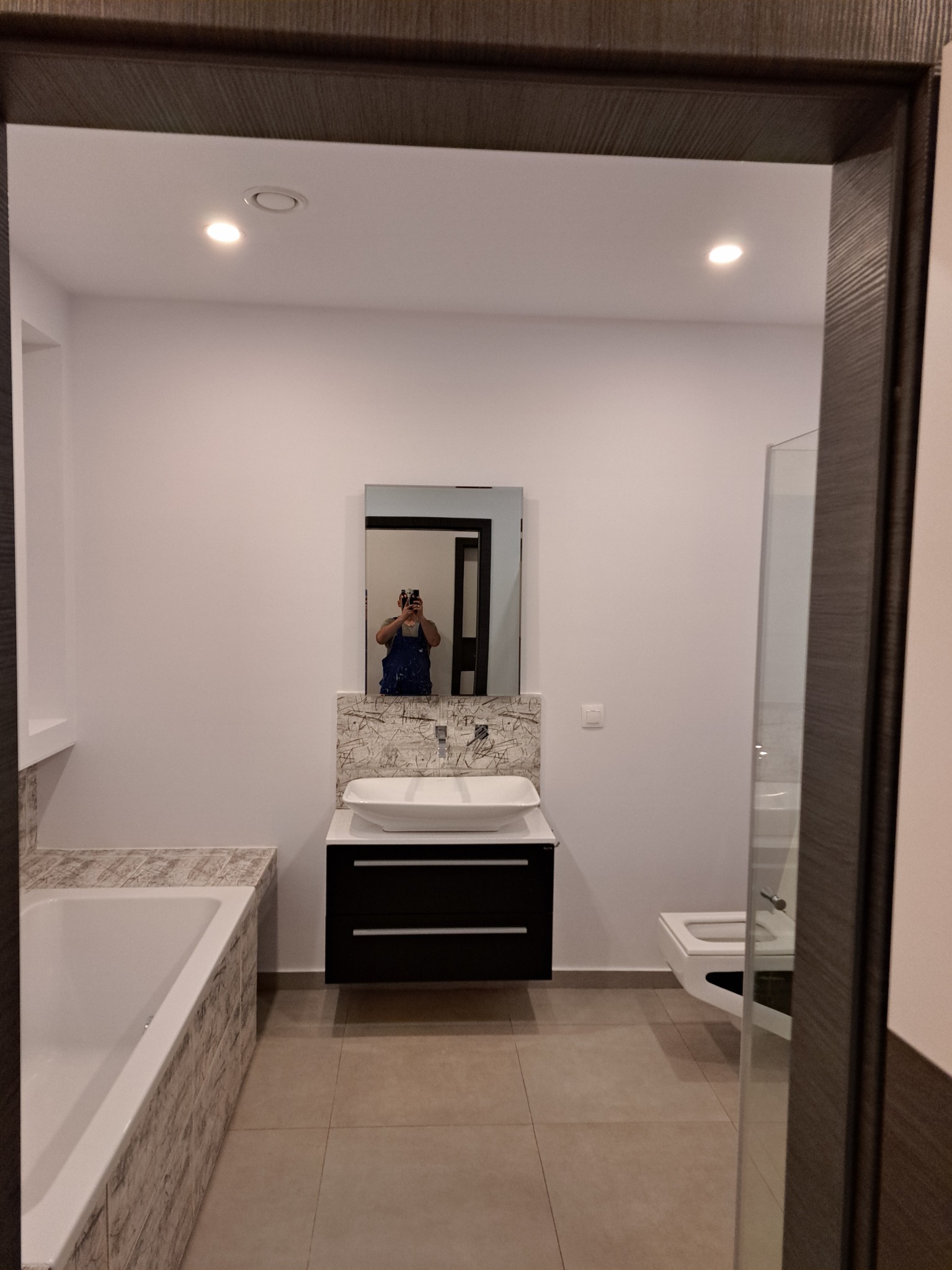 Nowoczesna, jasna łazienka z umywalką nablatową, czarną szafką pod umywalką, wanną obłożoną płytkami imitującymi kamień i toaletą w tle, widziana z perspektywy drzwi; w lustrze odbija się osoba...