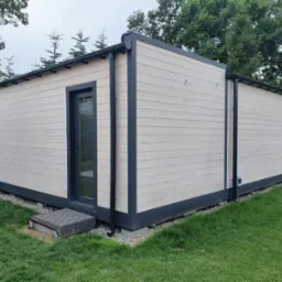 Zewnętrzny widok gotowego, parterowego domu z jasnego drewna z grafitowymi ramami okiennymi i obramowaniem, otoczonego zielonym trawnikiem i drzewami.