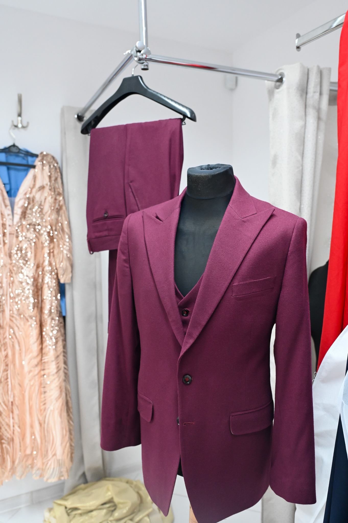 Burgundowy garnitur trzyczęściowy na manekinie, spodnie na wieszaku w tle, w pracowni krawieckiej, obok błyszcząca suknia wieczorowa.