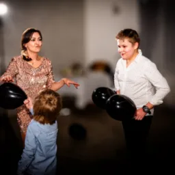 Kobieta w cekinowej sukience i dwójka dzieci z czarnymi balonami na tle rozproszonego światła podczas uroczystości.