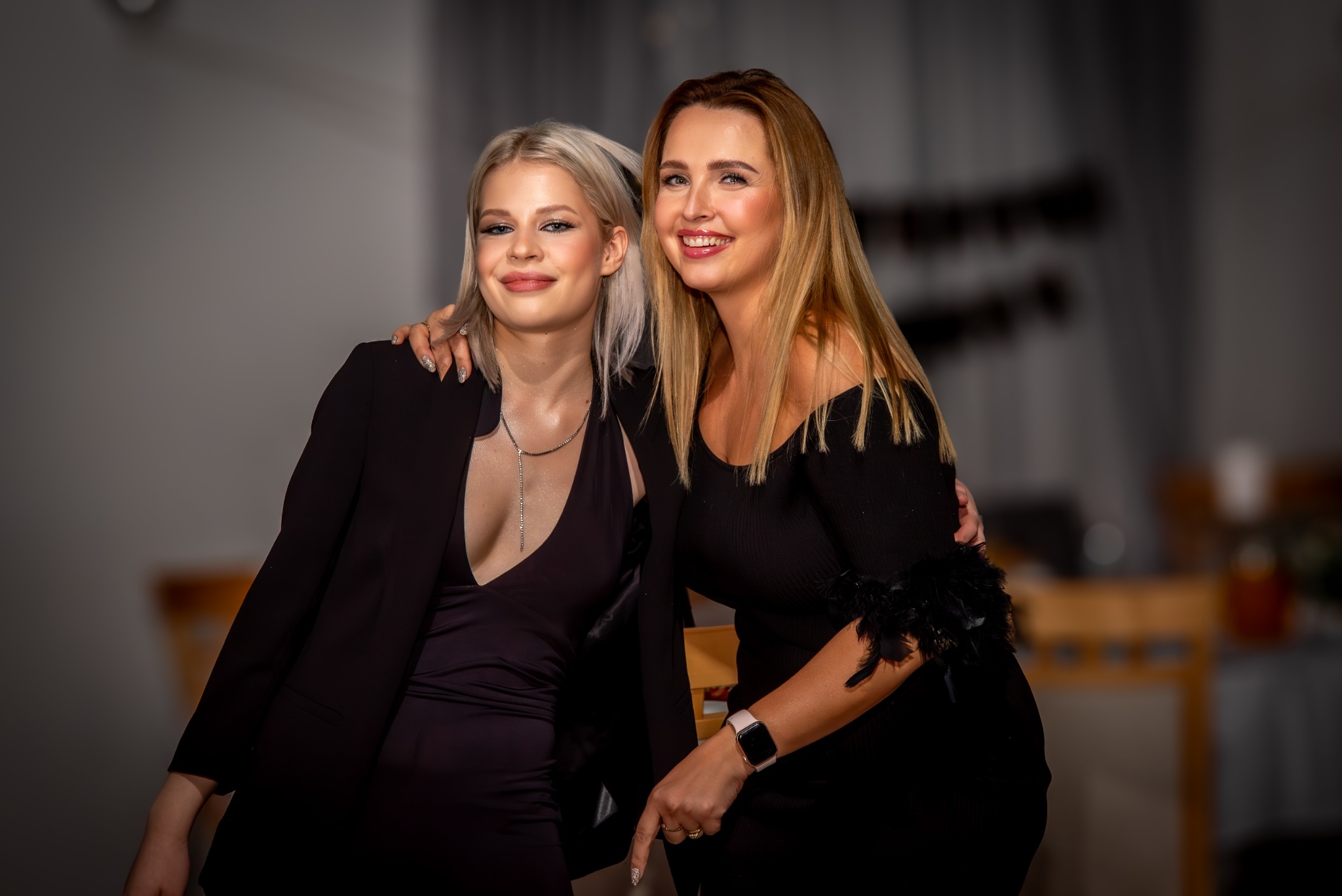 Dwie uśmiechnięte kobiety w czarnych sukienkach, blondynka i brunetka, pozują blisko siebie, widoczne detale: błyszczący manicure młodszej kobiety i pióra przy rękawie sukienki starszej.