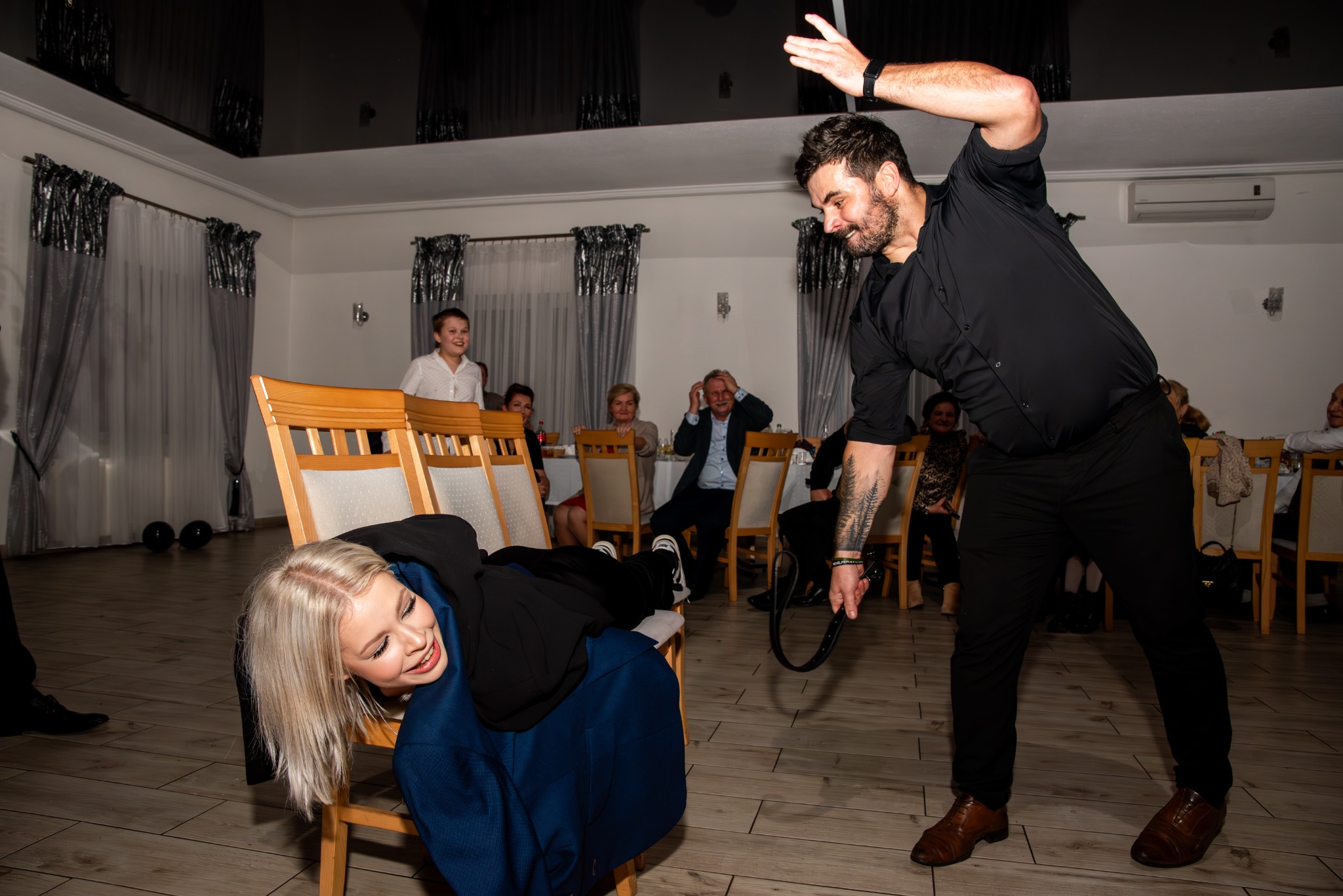 Zabawa weselna: kobieta leży na krześle, mężczyzna z paskiem w ręku gestykuluje, w tle goście weselni.
