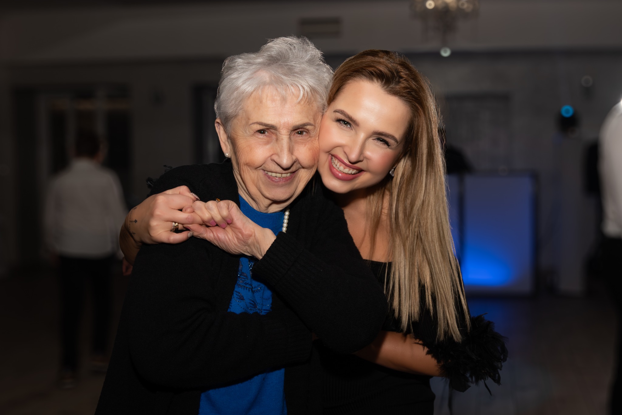 Uśmiechnięta młoda kobieta obejmuje starszą panią z perłami na szyi, w tle rozmyte postacie w sali bankietowej.