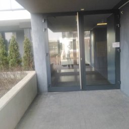 Euro Cleaning Olha Ivanenko - Szare, przeszklone drzwi wejściowe do budynku z widocznym odbiciem osoby w środku. Na zewnątrz wyłożony kostką chodnik i niewielkie drzewka iglaste.