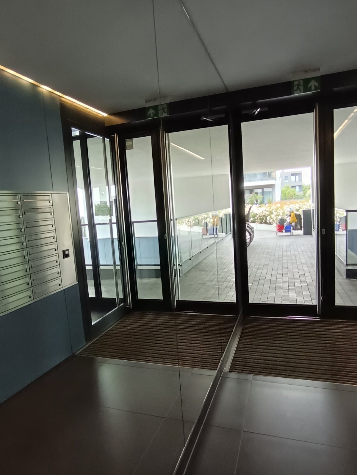 Wnętrze budynku z widokiem na zewnątrz przez szklane drzwi. Widoczne skrzynki pocztowe i wycieraczka. Na zewnątrz kostka brukowa, roślinność i elementy architektury.