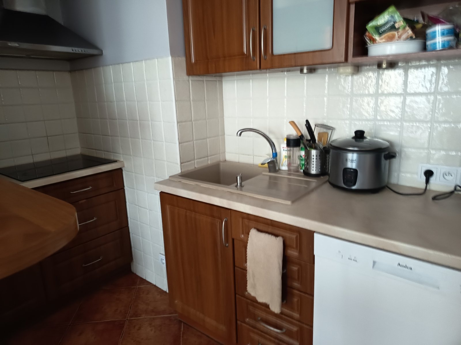 Umyta, czysta kuchnia z brązowymi szafkami, białymi płytkami i sprzętami AGD. Na blacie stoi srebrny garnek elektryczny i przybory kuchenne. W tle widać okap.