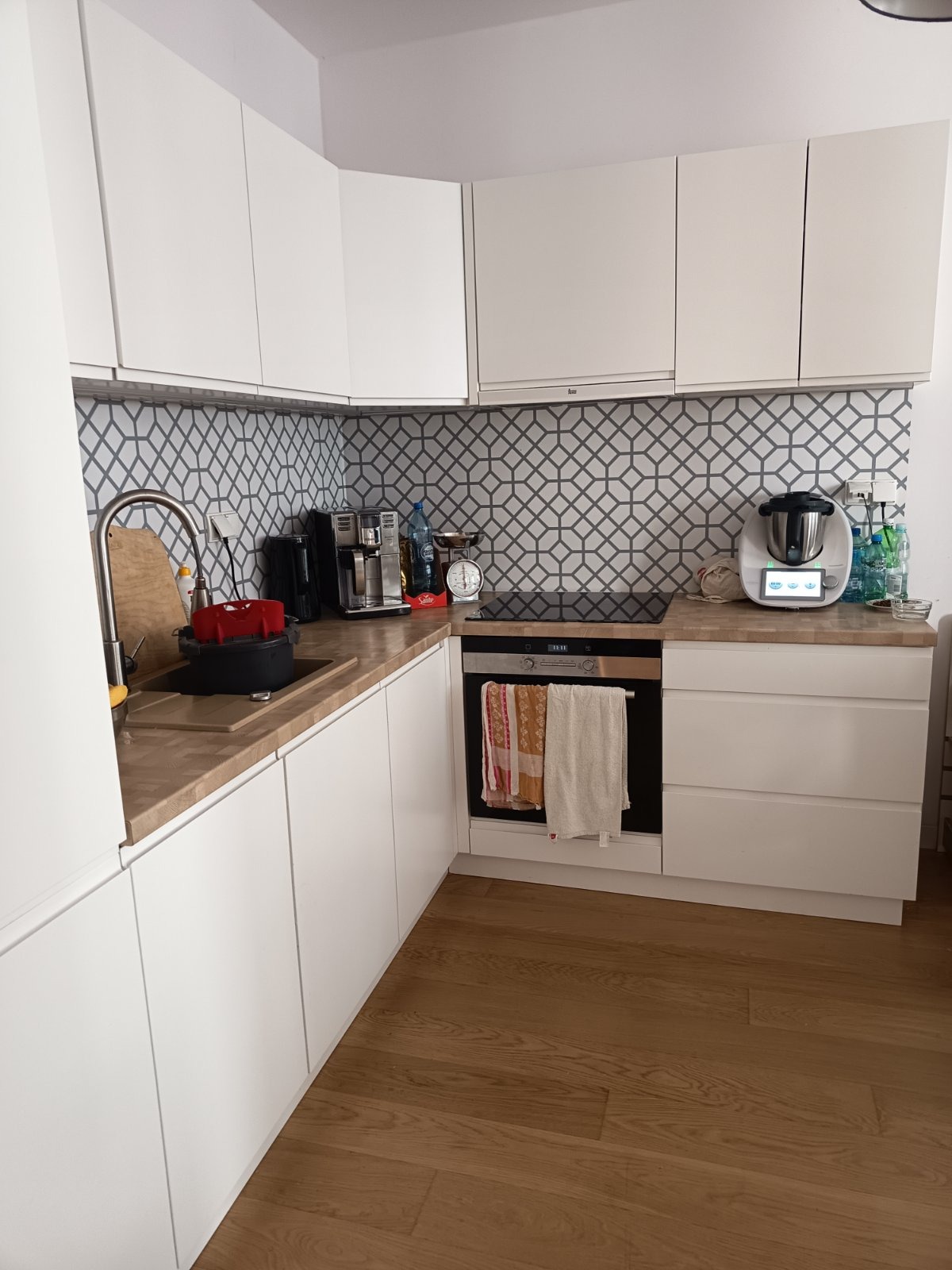 Jasna kuchnia z białymi szafkami, geometrycznym wzorem na ścianie, drewnianym blatem i podłogą. Widoczne naczynia w zlewie i sprzęty AGD na blacie.