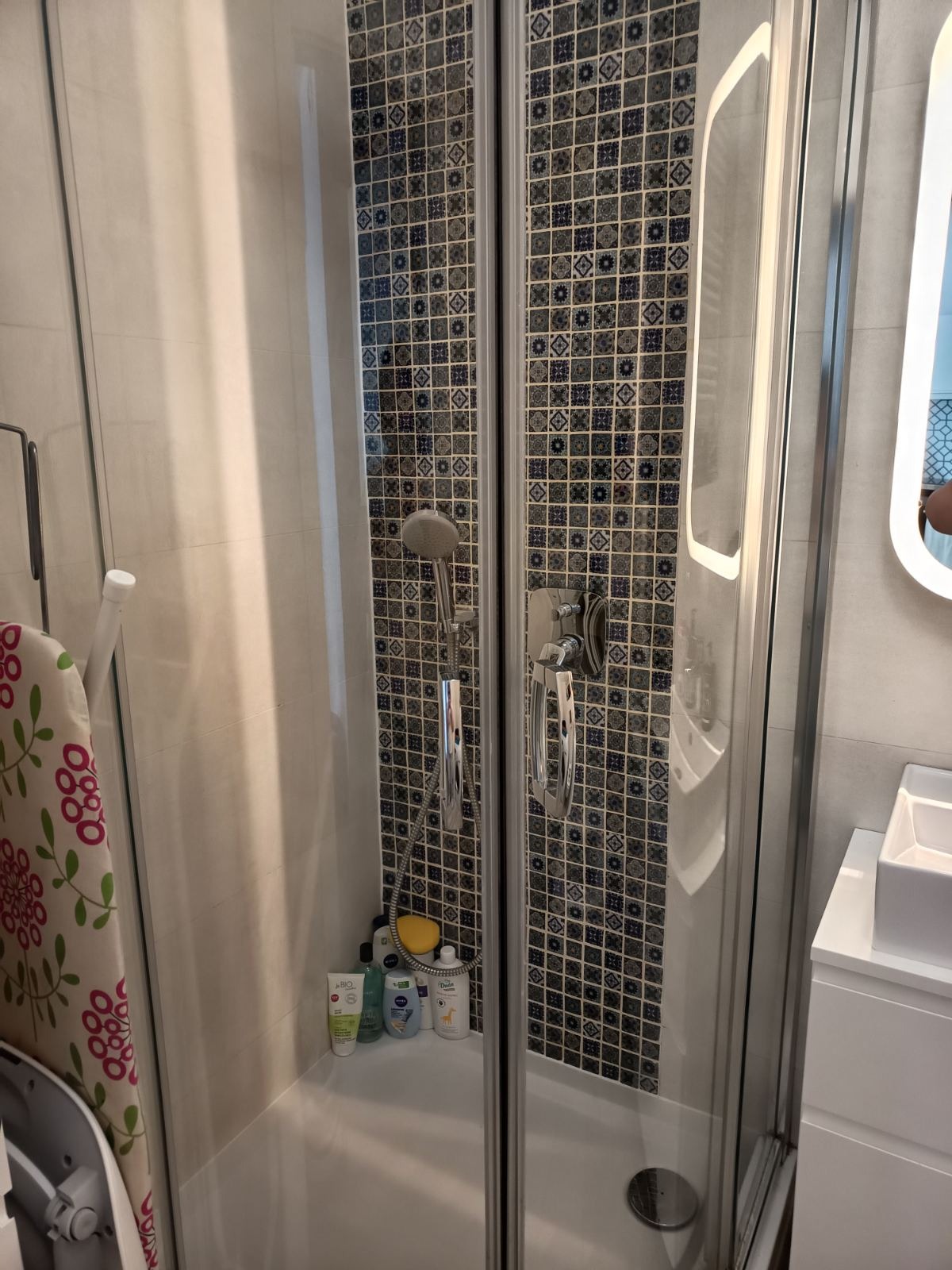 Kabina prysznicowa z dekoracyjnymi płytkami w łazience. Widoczne kosmetyki i suszarka na ręczniki. Biała ceramika łazienkowa. Ujęcie z bliska.