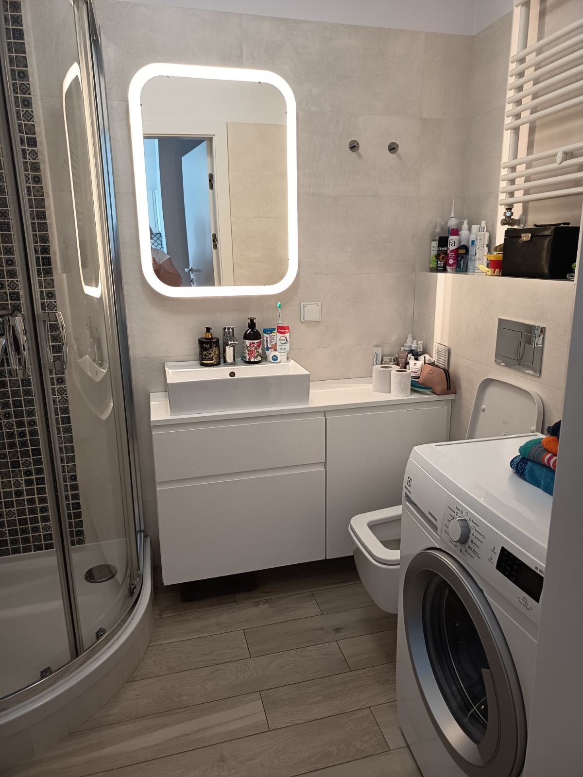 Jasna łazienka z kabiną prysznicową, umywalką z szafką, toaletą i pralką. Drewnopodobne płytki na podłodze. Lustro z podświetleniem LED. Minimalistyczny design.