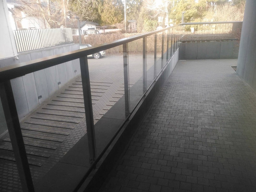 Nowoczesna balustrada ze szkła i metalu oddziela chodnik z kostki brukowej od rampy z drewnianymi elementami. W tle budynki i drzewa.