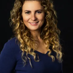 Portret uśmiechniętej kobiety z blond kręconymi włosami, ubrana w granatowy sweter na ciemnym tle.