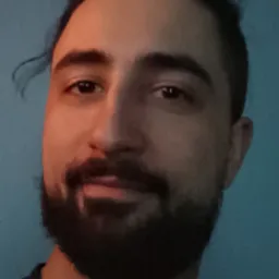 Portret mężczyzny z ciemną brodą i włosami związanymi z tyłu, na tle niebieskiej ściany, oświetlony z lewej strony.