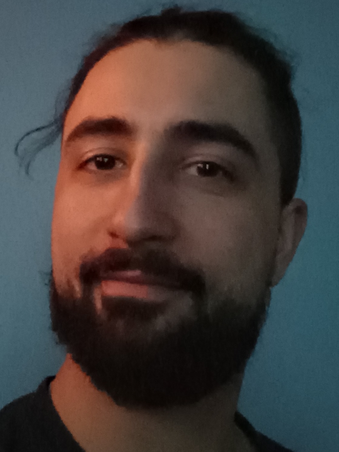 Portret mężczyzny z ciemną brodą i włosami związanymi z tyłu, na tle niebieskiej ściany, oświetlony z lewej strony.