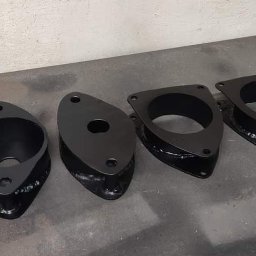 S-metal Siwiec Piotr - Cztery czarne, metalowe elementy konstrukcyjne z otworami, prawdopodobnie wsporniki lub łączniki, leżące na szorstkiej, metalowej powierzchni.