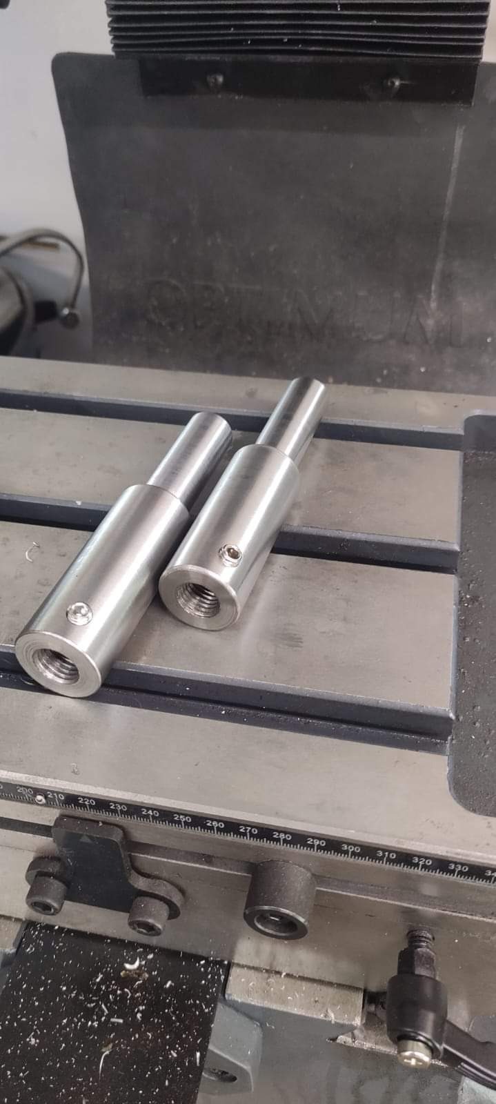 Dwa metalowe adaptery z gwintem wewnętrznym i śrubą ustalającą leżące na stole frezarki. Widoczne wióry po obróbce.